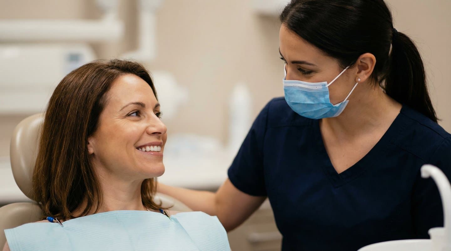 A patient in a dental chair smiling while a clinician in scrubs and mask rests a reassuring hand on her shoulder