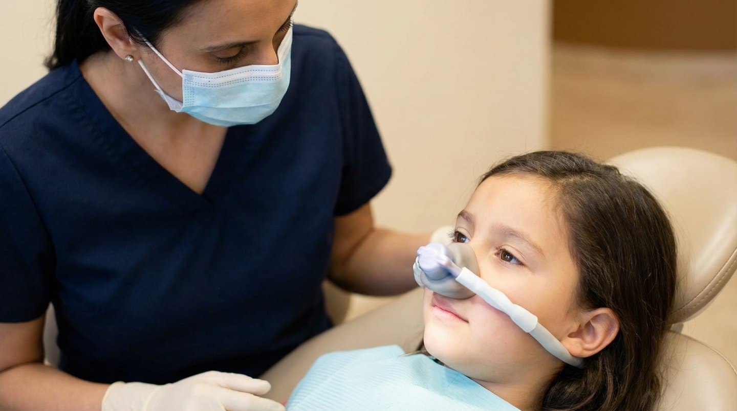 A relaxed child in a dental chair wearing a nitrous oxide nose mask while a dental professional in scrubs provides care